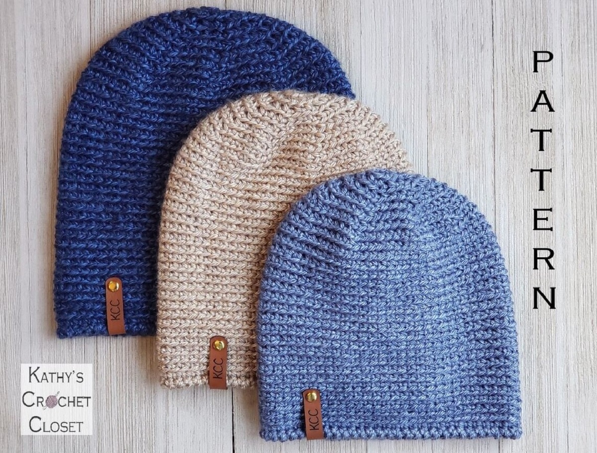 Pale blue, cream, and dark blue beanie stacked on top of each other with a tan leather tag on the bottom left on a pale wooden background. 
