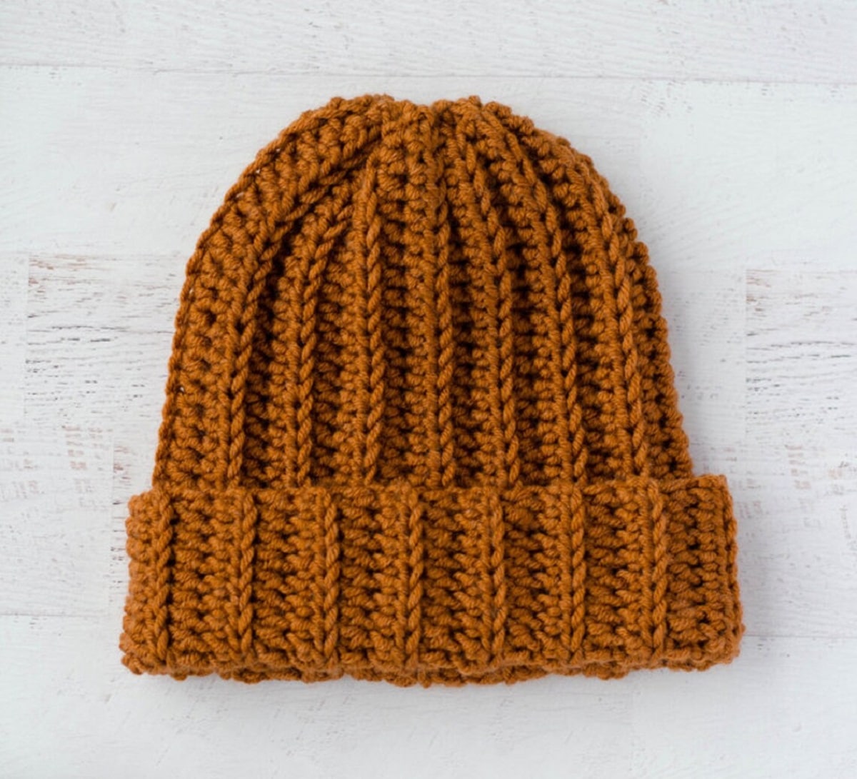 A dark orange ribbed crochet beanie with a thick banding on the bottom on a white wooden background.