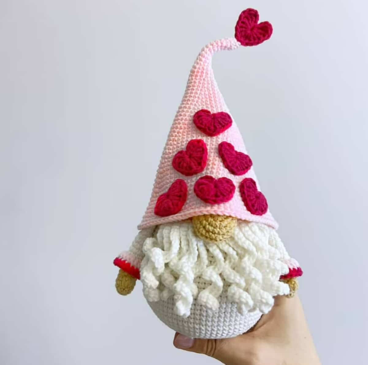 A hand holding a crochet stuffed gnome wearing a large pale pink pointed hat with pink flowers stitched all over and a white beard sticking out.Valentine Gnome Crochet Pattern - (HappyDollsHandMade)