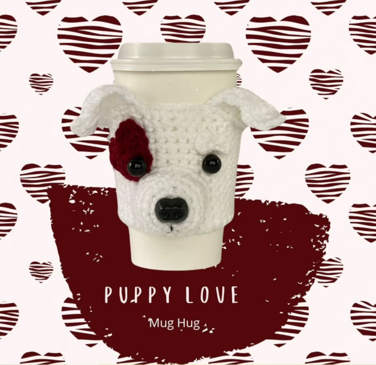 A white travel coffee cup with a crochet dog warmer over the cup. The white dog has ears stitching out and a red heart over one eye.